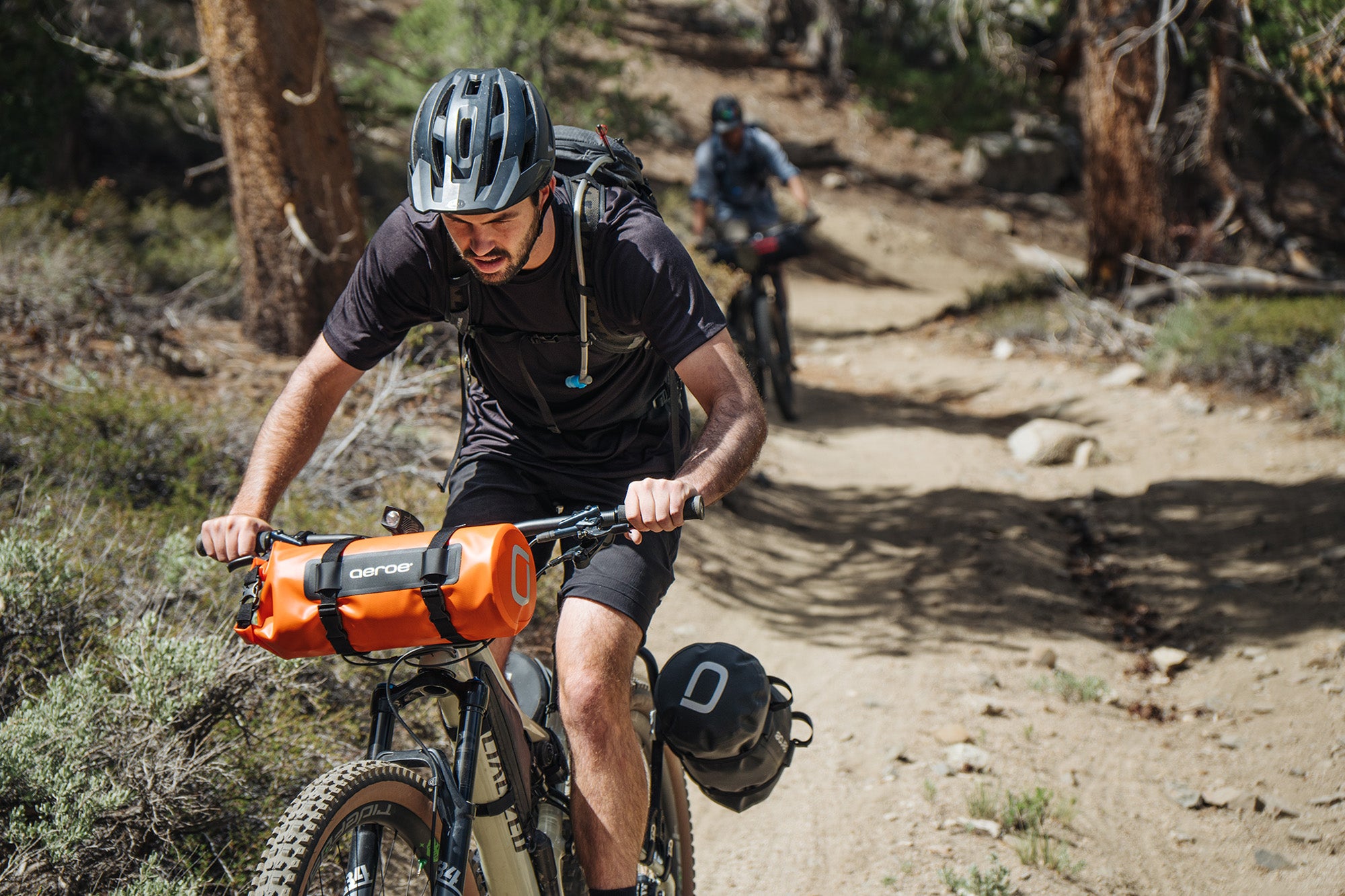 Man riding a mountain bike on a trail with a backpack and orange dry bag
